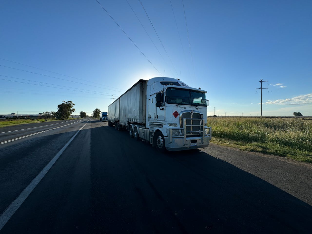 Home A silver semi truck driving on an open highway under a clear blue sky.