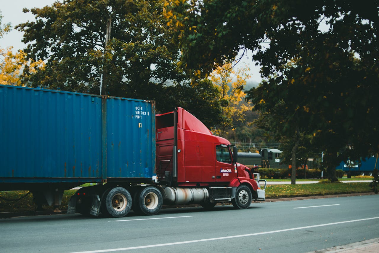 Home A red truck transports a blue cargo container on a scenic, tree-lined road.